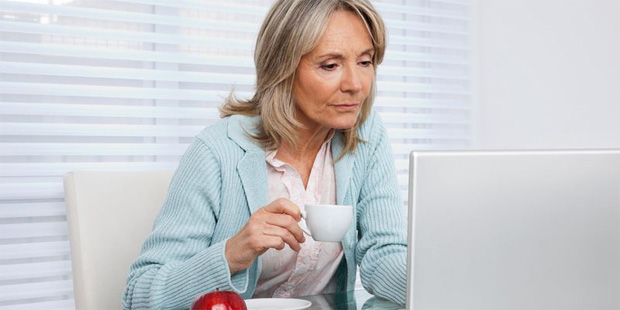 Пожилая женщина с чашкой кофе сидит перед ноутбуком